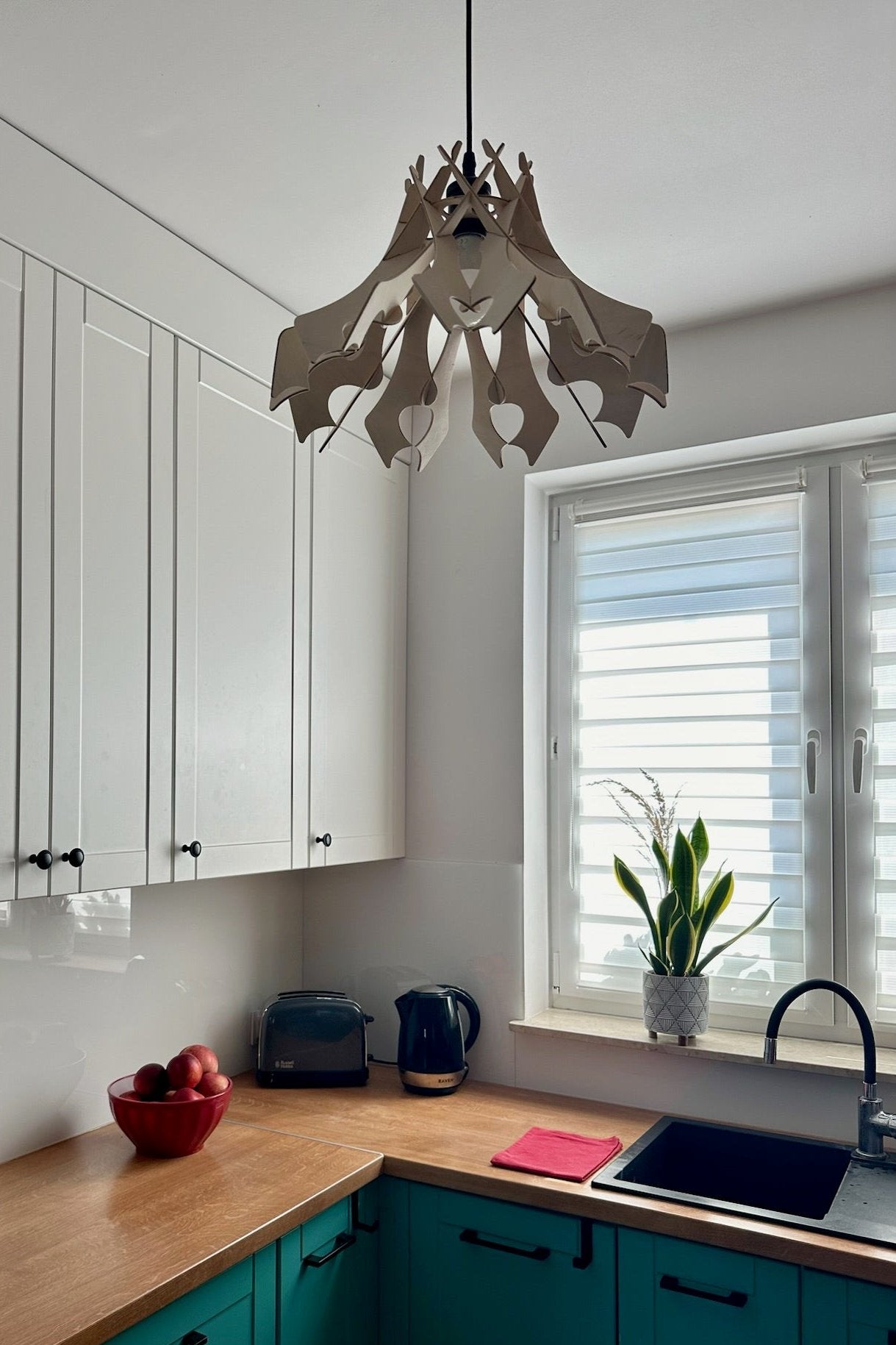 Natural birch wood pendant lampshade with a unique, curved design, hanging in a modern kitchen. The lamp is turned off, showcasing its intricate shape against the clean white cabinets, adding an elegant touch to the minimalist space.