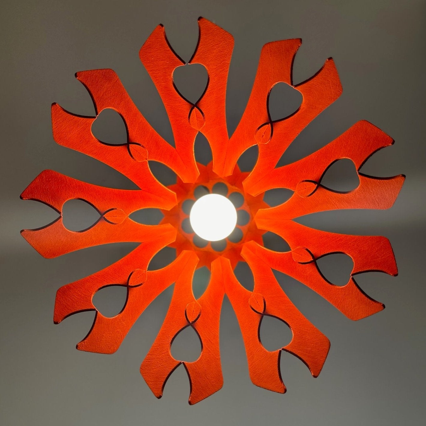 Bottom view of a scarlet-red pendant lampshade with intricate heart-shaped cutouts, creating a floral pattern of light on the ceiling. The bright light source is centered, enhancing the symmetrical design.