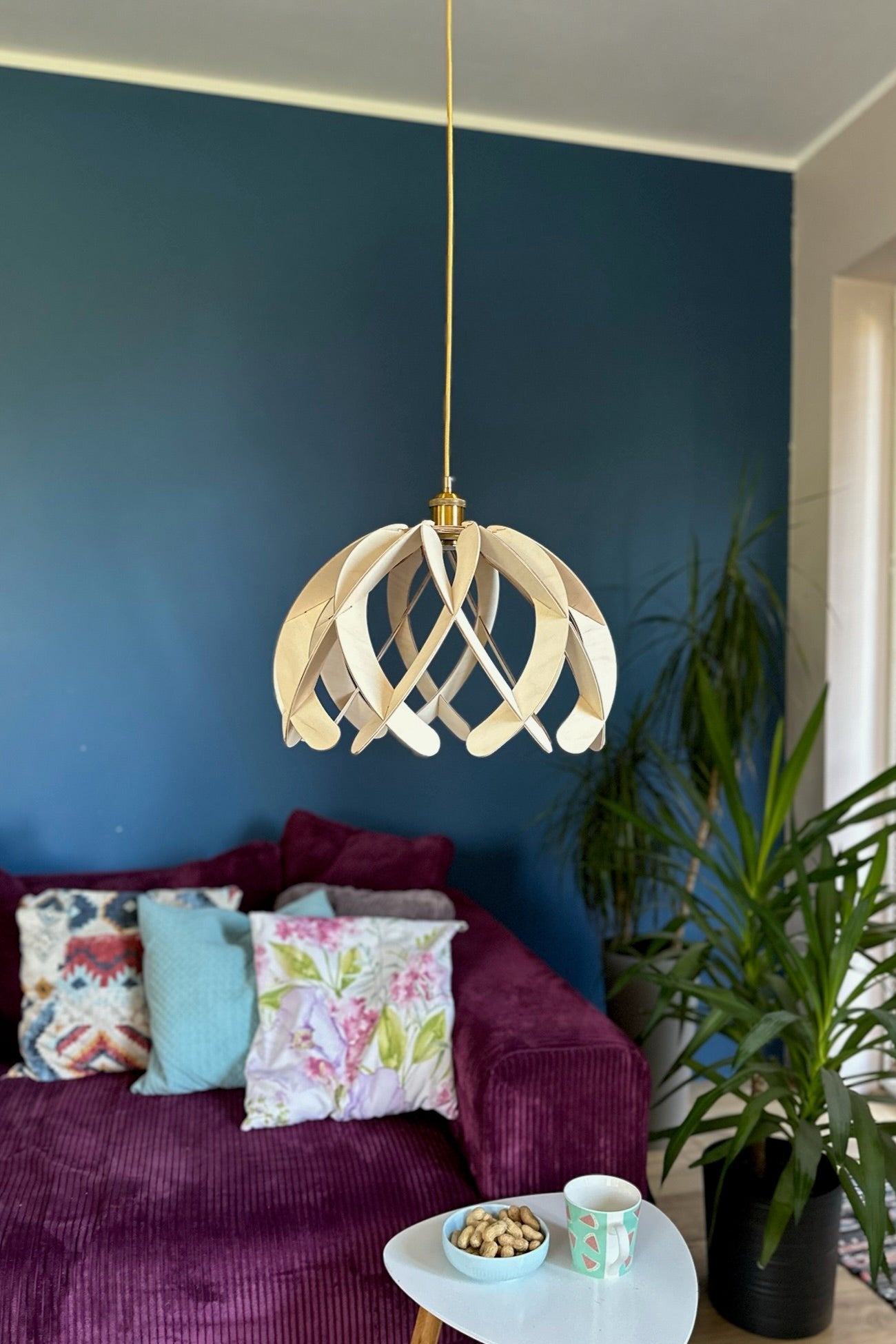 Natural birch wood pendant lampshade with a minimalist, rounded design hanging above a cozy living room setup. The gold cord complements the modern and rustic décor of the room, including a purple sofa and vibrant pillows.